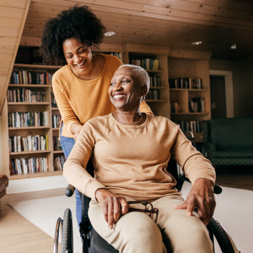 Older woman in a wheelchair smiling; younger woman standing behind her, both looking happy.