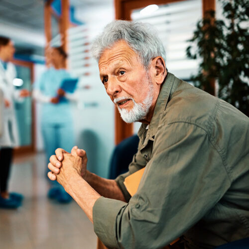 Pensive man with gray hair sitting, medical staff in the background.