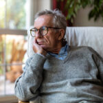 Elderly man sitting indoors, looking out the window with a thoughtful expression, symbolizing concern or reflection.