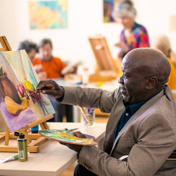 Man in a wheelchair painting a canvas with colorful flowers, in a bright art class setting.