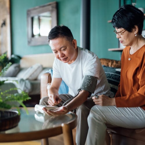 Senior Asian couple checking blood pressure at home. Wife examining blood pressure on her husband's arm with a blood pressure monitor. Elderly and healthcare concept