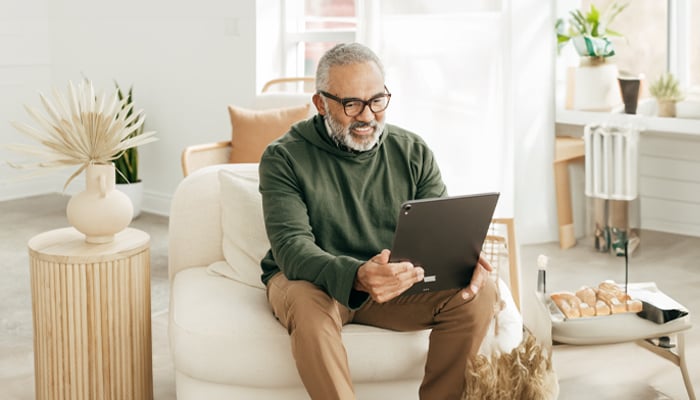 Older man sitting on a sofa and smiling while using a tablet for online communication or telehealth services.”